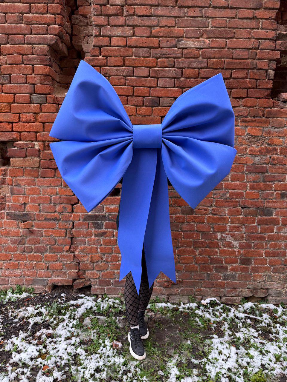 May include: A large, vibrant blue bow with long, flowing tails. The bow is the focal point, set against a weathered brick wall. The bow's size and color make it a striking decorative element.