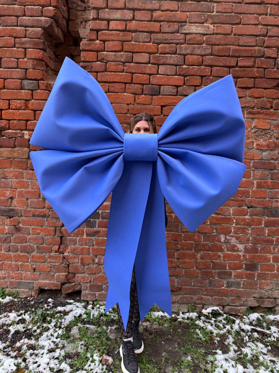 May include: A large, vibrant blue bow, possibly for decoration or a prop, dominates the frame. The bow is held in front of a person, obscuring their face. The background is a weathered brick wall, with patches of snow and greenery at the base.