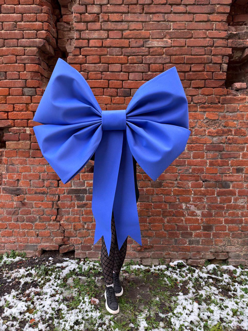 May include: A large, vibrant blue bow with long streamers. The bow is the focal point, set against a weathered red brick wall. The bow's size and color create a striking visual contrast.