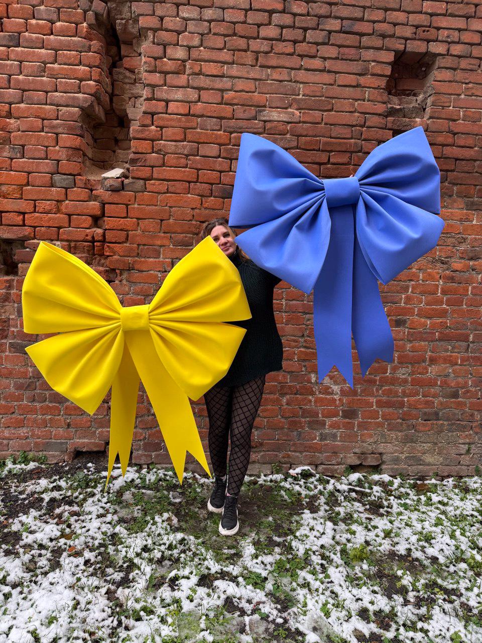 May include: A person holds two large decorative bows. One bow is bright yellow, and the other is a vibrant blue. The bows are made of a smooth material and have long, flowing tails. The background is a weathered brick wall.