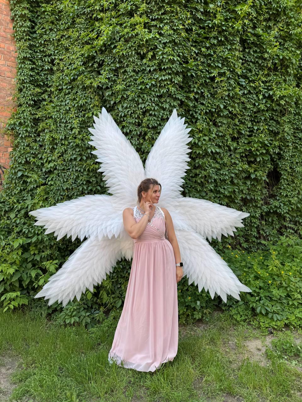 May include: A woman wearing a light pink dress and large white angel wings stands in front of a wall covered in green foliage. The wings are made of white feathers and are spread out behind the person.