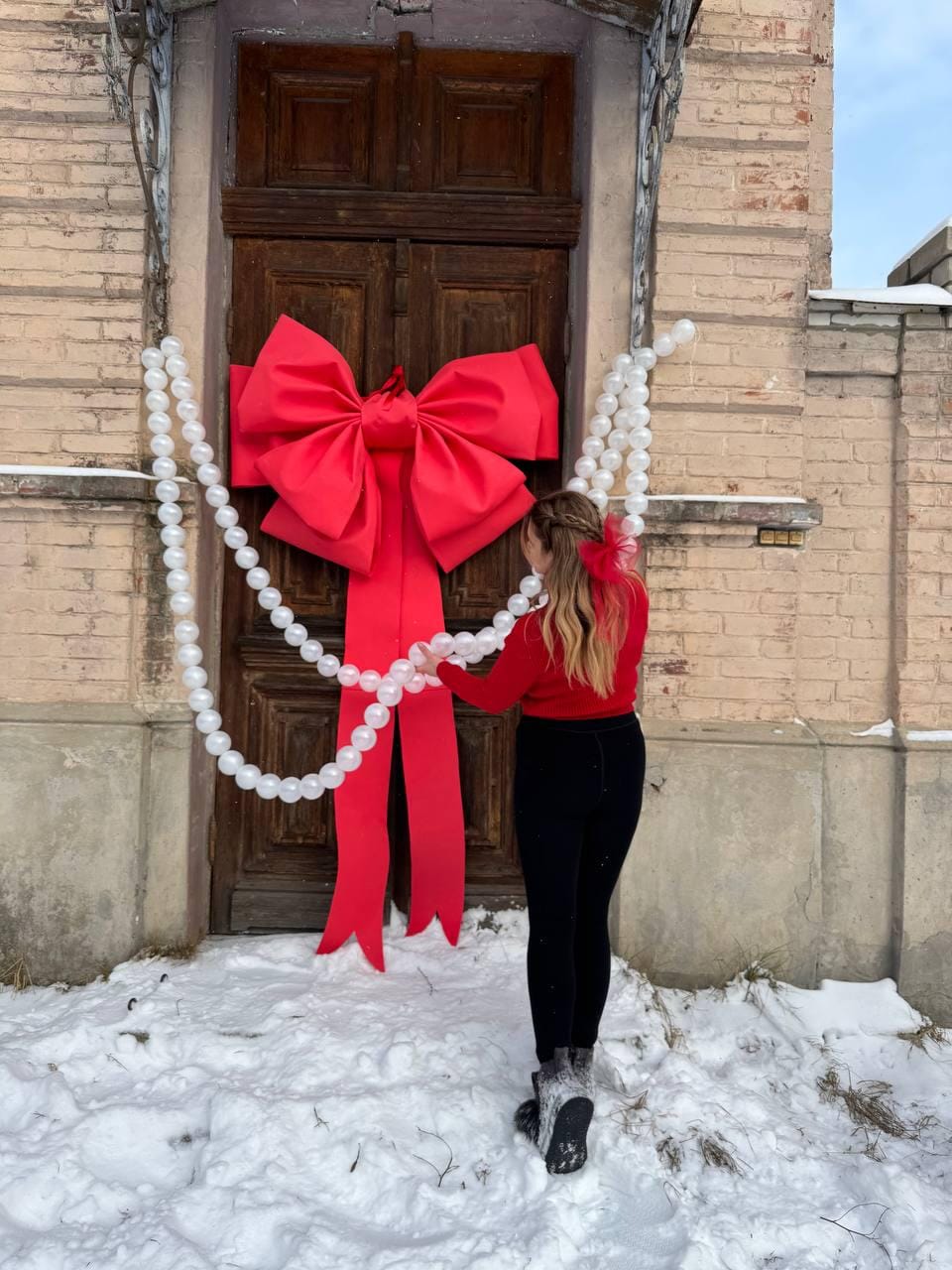 May include: A large red bow adorns a wooden door, with long flowing ribbons. A strand of white, round decorations hangs on the side. The scene is set outdoors with snow on the ground, suggesting a festive or holiday theme.