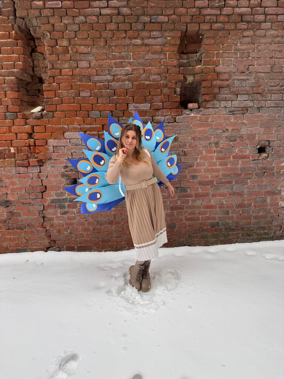 May include: A woman wearing a tan dress and brown boots stands in the snow, posing in front of a brick wall. She is wearing a large, colorful peacock feather costume with blue, orange, and white accents.