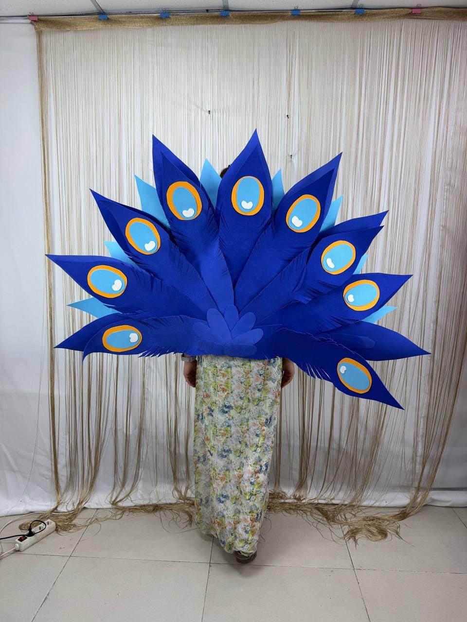 May include: A large, vibrant blue peacock feather costume. The costume features large, pointed feathers with oval-shaped eye designs in shades of blue, yellow, and white. The costume is worn by a person wearing a floral print dress.