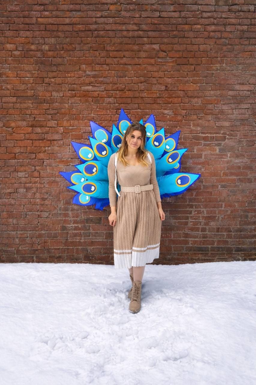 May include: A woman stands in front of a brick wall with a large, blue and turquoise peacock feather decoration. She wears a tan dress with a matching belt and boots. The ground is covered in snow.
