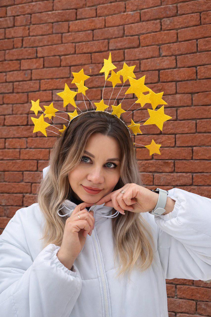 May include: A yellow starburst crown with glittery stars of varying sizes. The celestial headpiece is attached to a silver headband. The person in the image is wearing a white jacket and is in front of a brick wall.