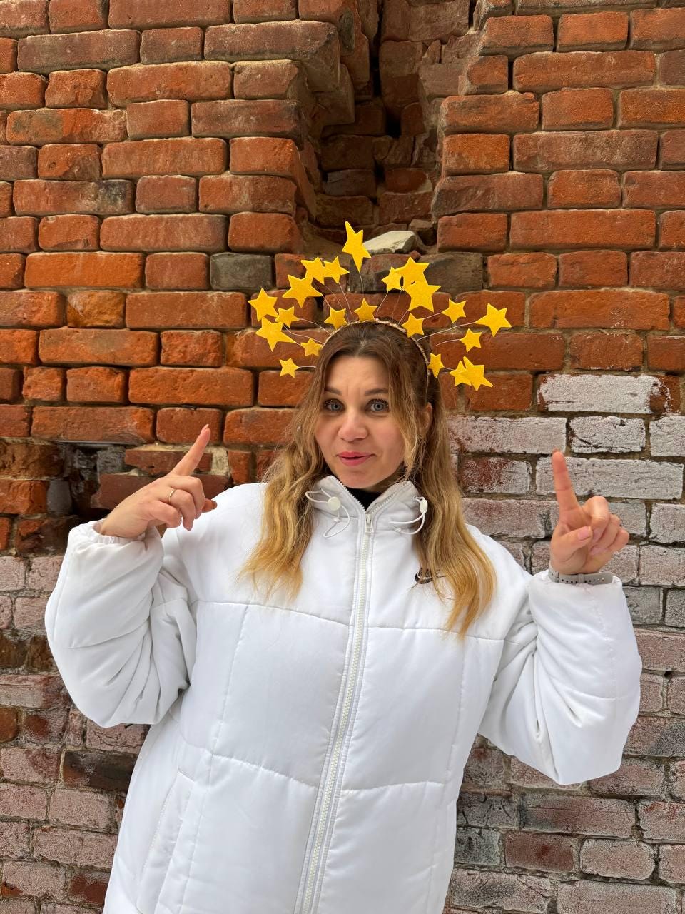 May include: A yellow starburst crown is worn by a person in a white puffer jacket. The crown is made of yellow stars attached to a headband. The background is a weathered brick wall.