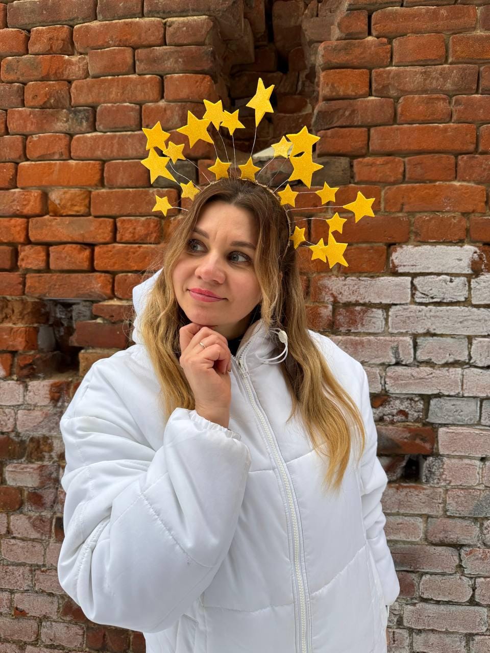 May include: A gold-toned headband adorned with numerous yellow star-shaped ornaments. The stars are arranged in a halo-like fashion. The person in the image is wearing a white jacket. The background is a weathered brick wall.