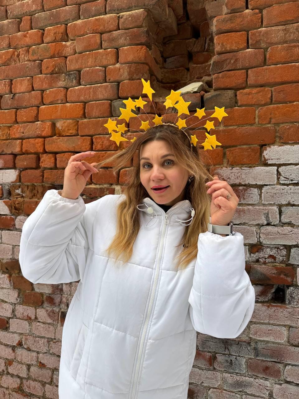 May include: A yellow star-shaped headband is worn by a person wearing a white puffer jacket. The headband has multiple star shapes attached to a wire frame. The person is standing in front of a red brick wall.