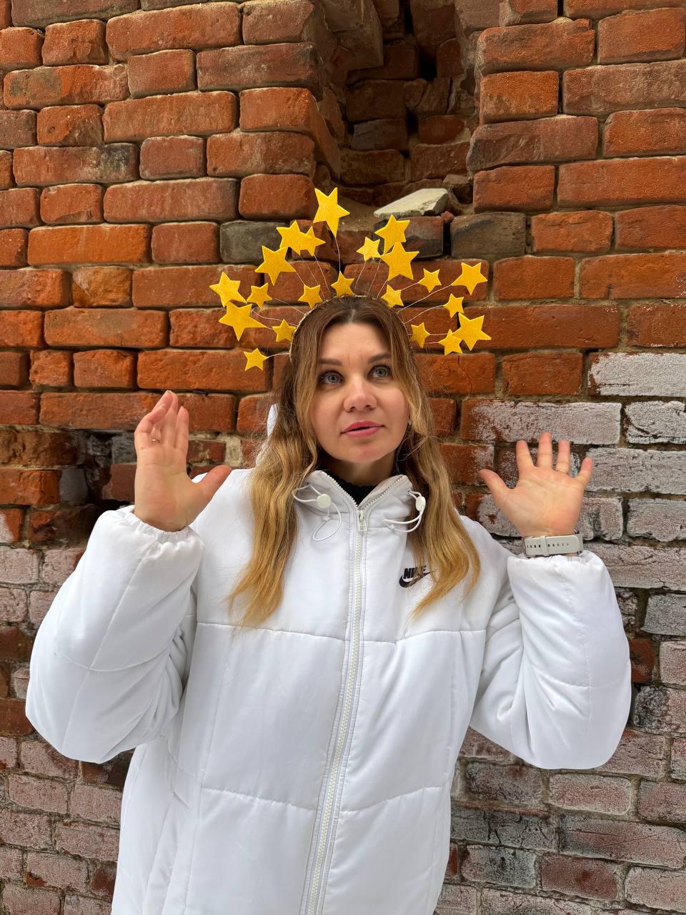 May include: A woman wearing a yellow star-shaped headpiece and a white puffer jacket with a Nike logo. The background is a weathered brick wall.