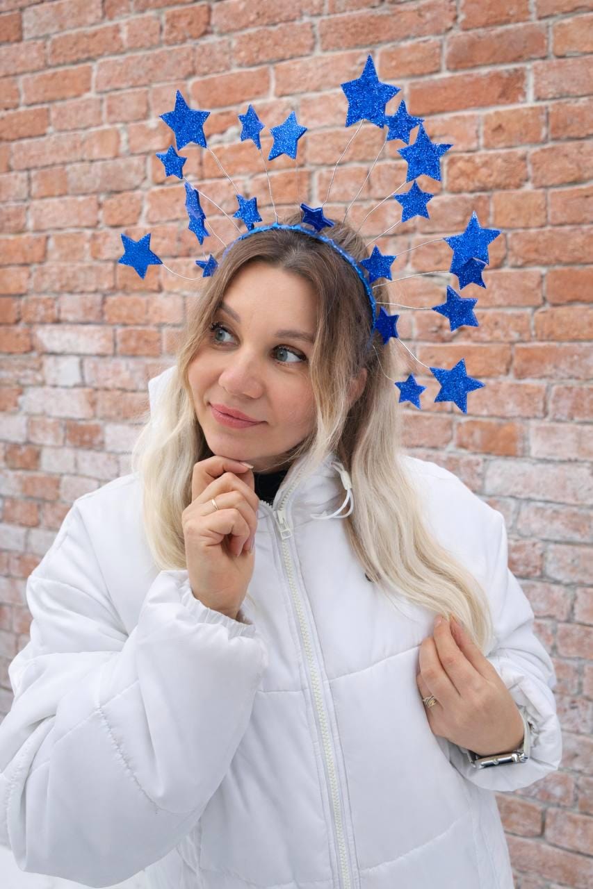 May include: A blue starburst headband with glittery stars. The headband is worn by a person wearing a white puffer jacket. The background is a brick wall.
