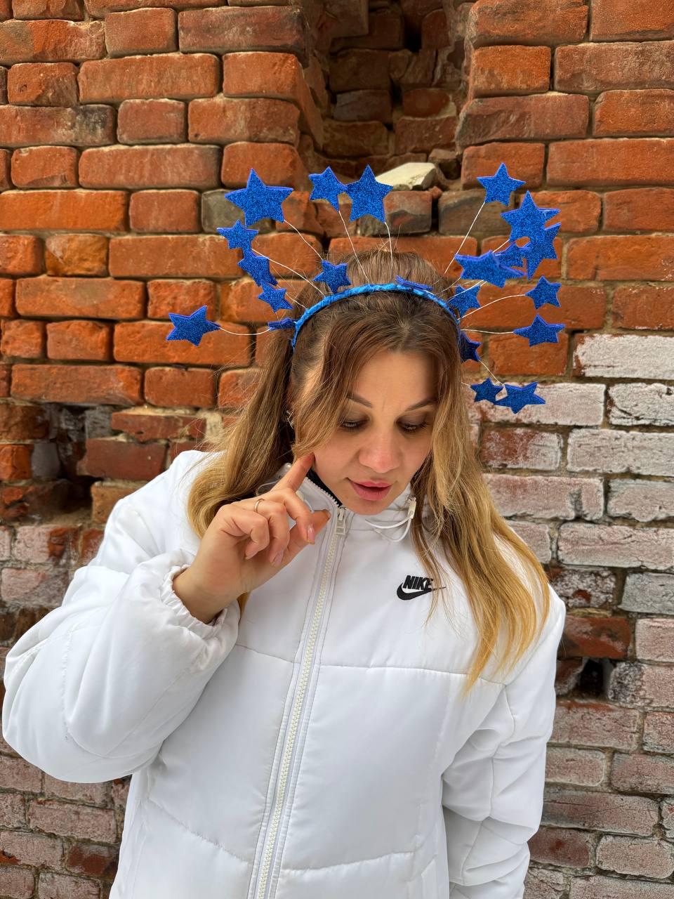 May include: A blue star-shaped headband with multiple stars attached to a wire frame. The headband is worn by a person wearing a white jacket with a Nike logo. The background is a brick wall.