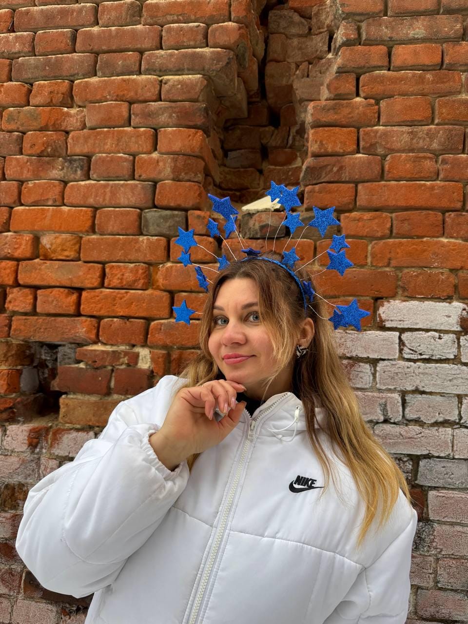 May include: A woman wearing a blue headband adorned with glittery blue stars. She is wearing a white puffer jacket with a black Nike logo. The background is a weathered brick wall.
