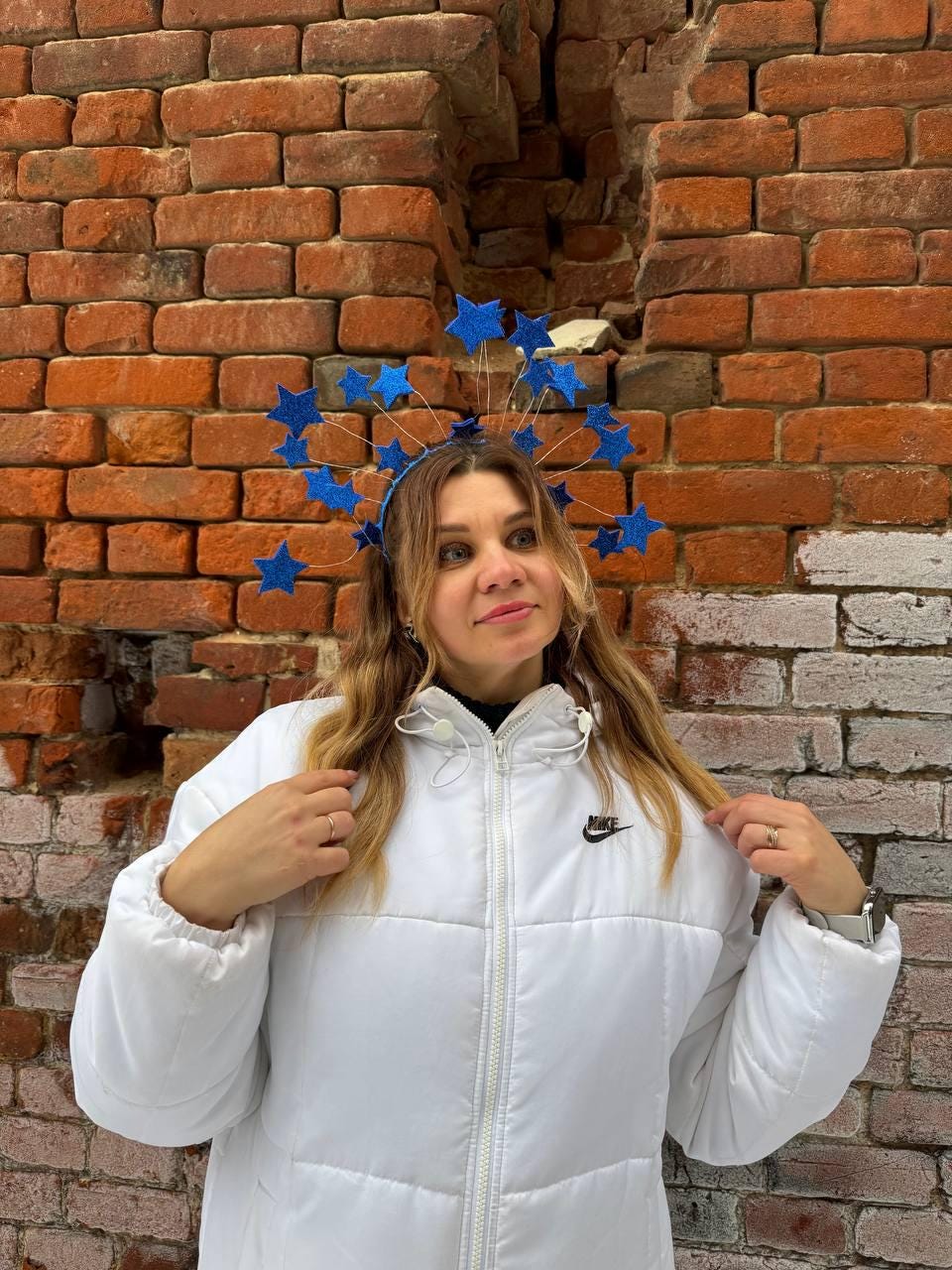 May include: A blue star-shaped headpiece with a matching headband. The headpiece is worn over long, wavy blonde hair. The person is wearing a white puffer jacket with a Nike logo on the chest, standing in front of a brick wall.