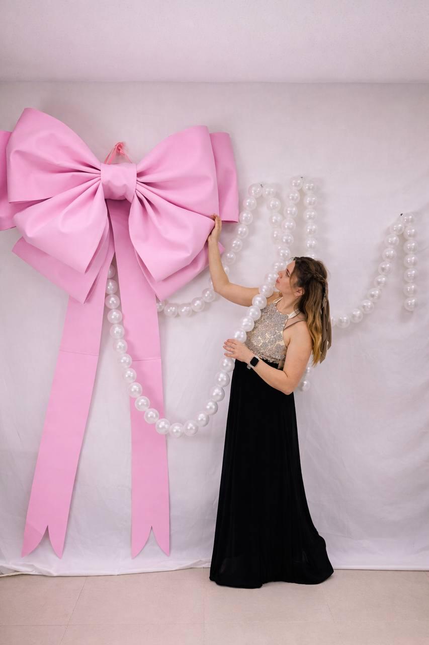 May include: A large, pink bow with long streamers is the focal point. A woman in a black dress and a beaded top is holding a strand of white pearls. The bow is mounted on a white backdrop.