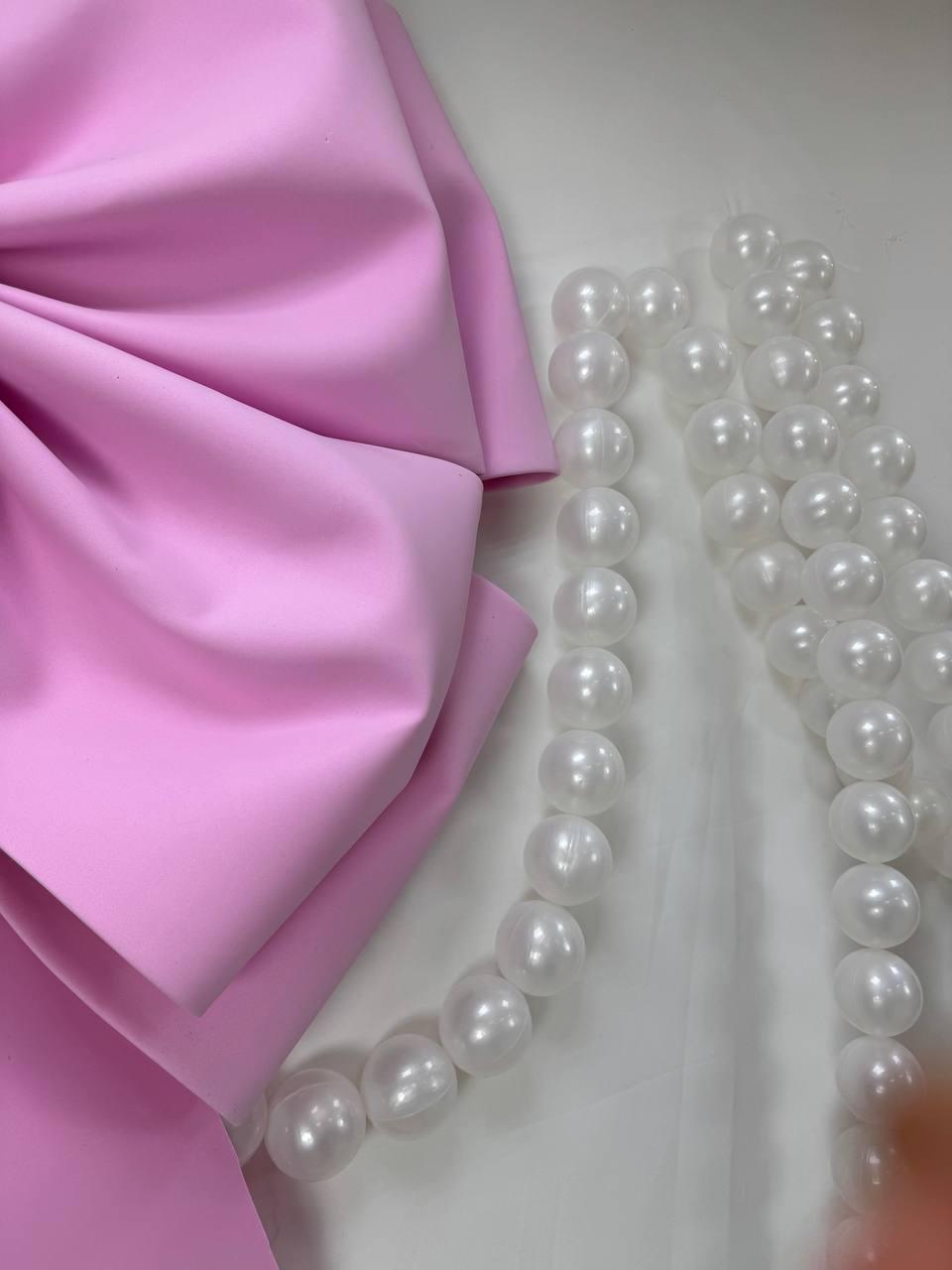 May include: A large, light pink fabric bow is positioned next to two strands of white, round beads. The bow has a smooth texture and is folded in multiple layers. The beads are evenly spaced and appear to be made of a glossy material.