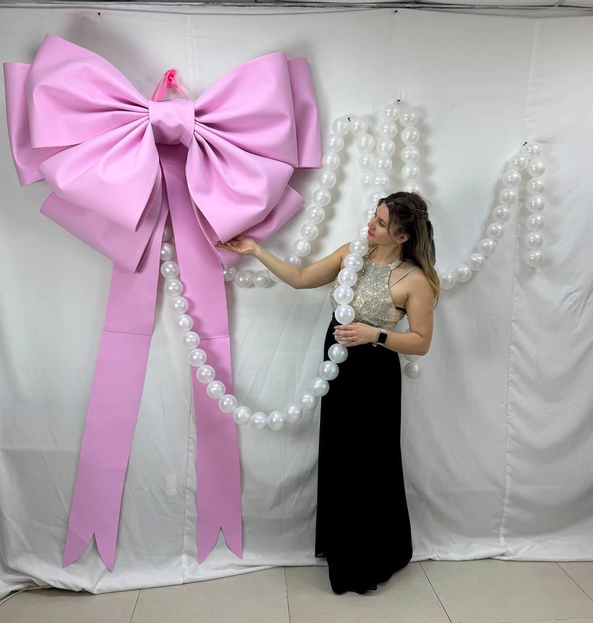 May include: A large, light pink bow is mounted on a white wall. The bow is paired with a string of white pearls. The bow's long tails hang down. The bow is a decorative element, possibly for a party or event.
