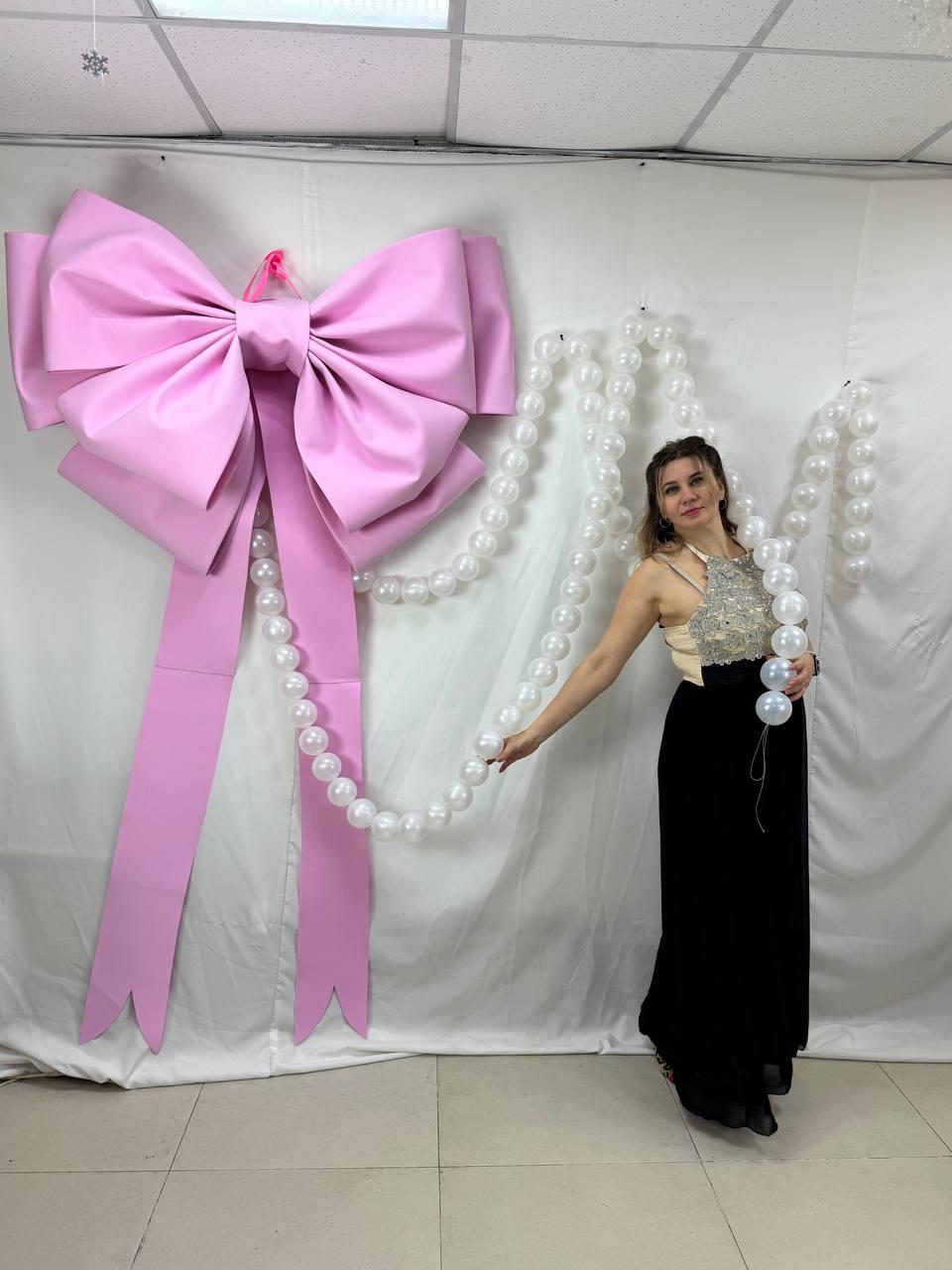 May include: A large, pink bow with long streamers hangs on a white backdrop. A strand of white pearls is draped near the bow. A person is holding a strand of white pearls and wearing a black skirt and a cream-colored top.
