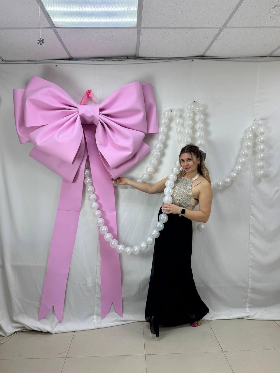 May include: A large, pink bow decoration with long streamers. A woman is holding a strand of white pearls. The bow is mounted on a white backdrop, creating a festive and elegant display.