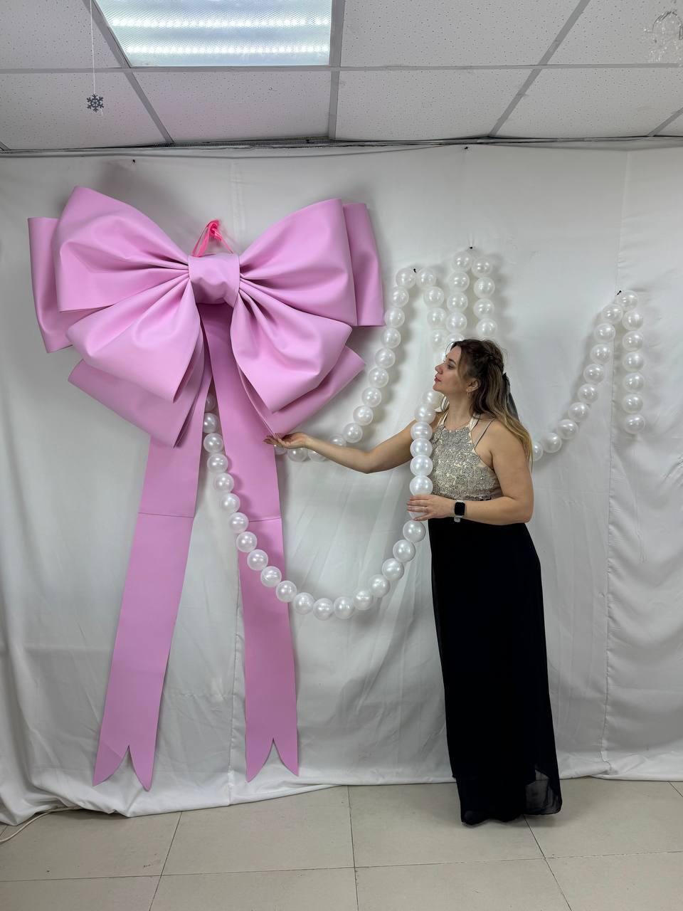 May include: A large, pink bow with long streamers hangs on a white wall. The bow is made of a smooth material and is the focal point of the image. A strand of large, white pearls is draped nearby. The bow is a decorative element.