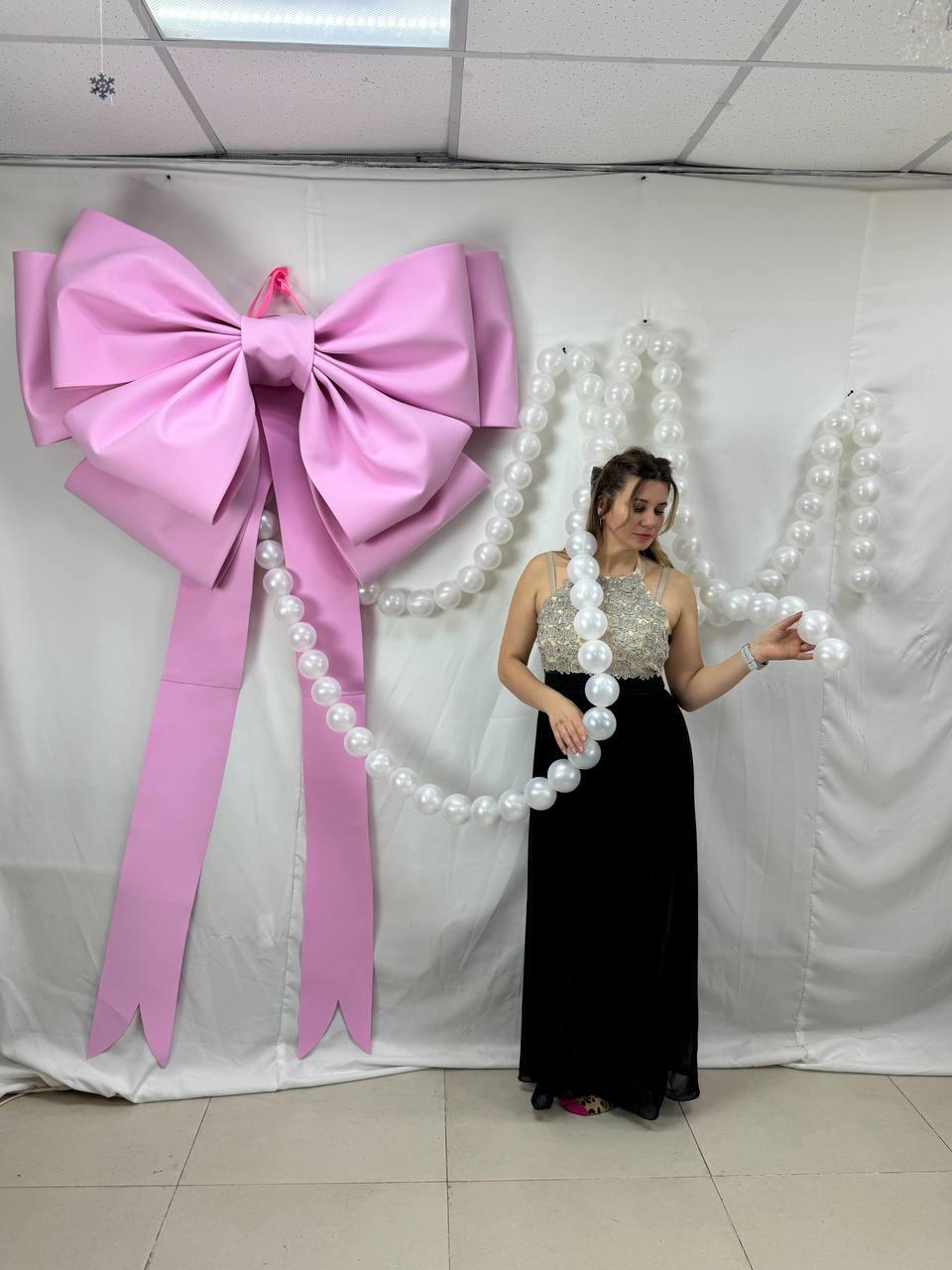 May include: A large, pink bow decoration with long streamers hangs on a white wall. A strand of white pearls is draped nearby. A person wearing a black dress and a beaded top is standing next to the pearls.