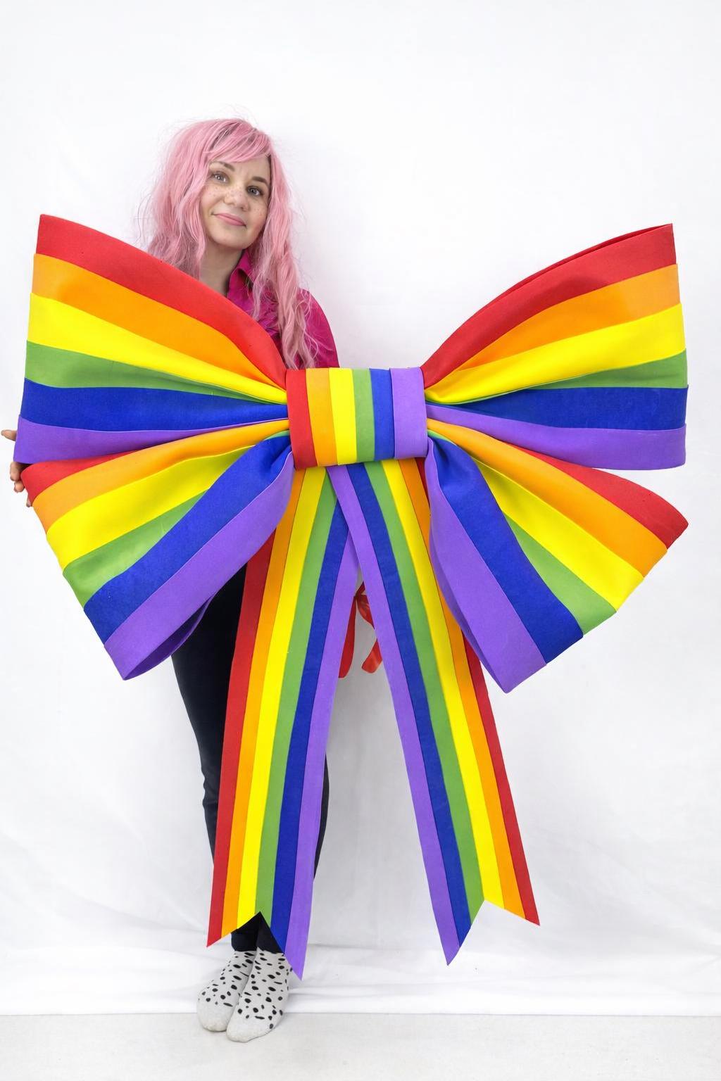 May include: A large, colorful rainbow bow with red, orange, yellow, green, blue, and purple stripes. The bow is held up against a white background. The bow is a decorative item.