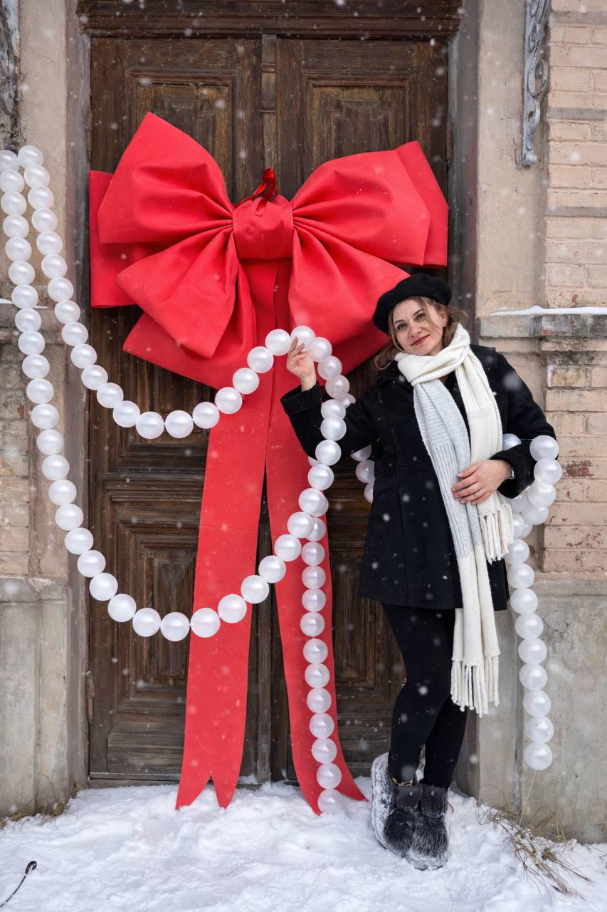 May include: A large red bow and matching ribbons adorn a wooden door, complemented by a string of white, round ornaments. A person stands nearby, wearing a black coat, hat, and scarf, with snow on the ground.