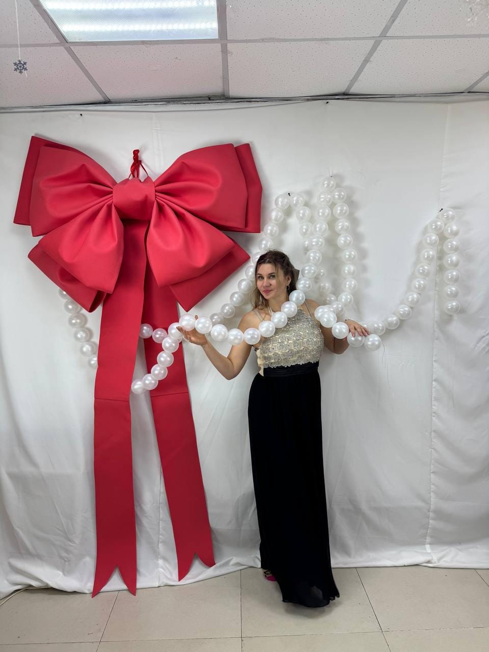 May include: A large, bright red bow with long streamers hangs on a white backdrop. A woman in a sequined top and black skirt holds a strand of white pearls. The bow is a festive decoration.