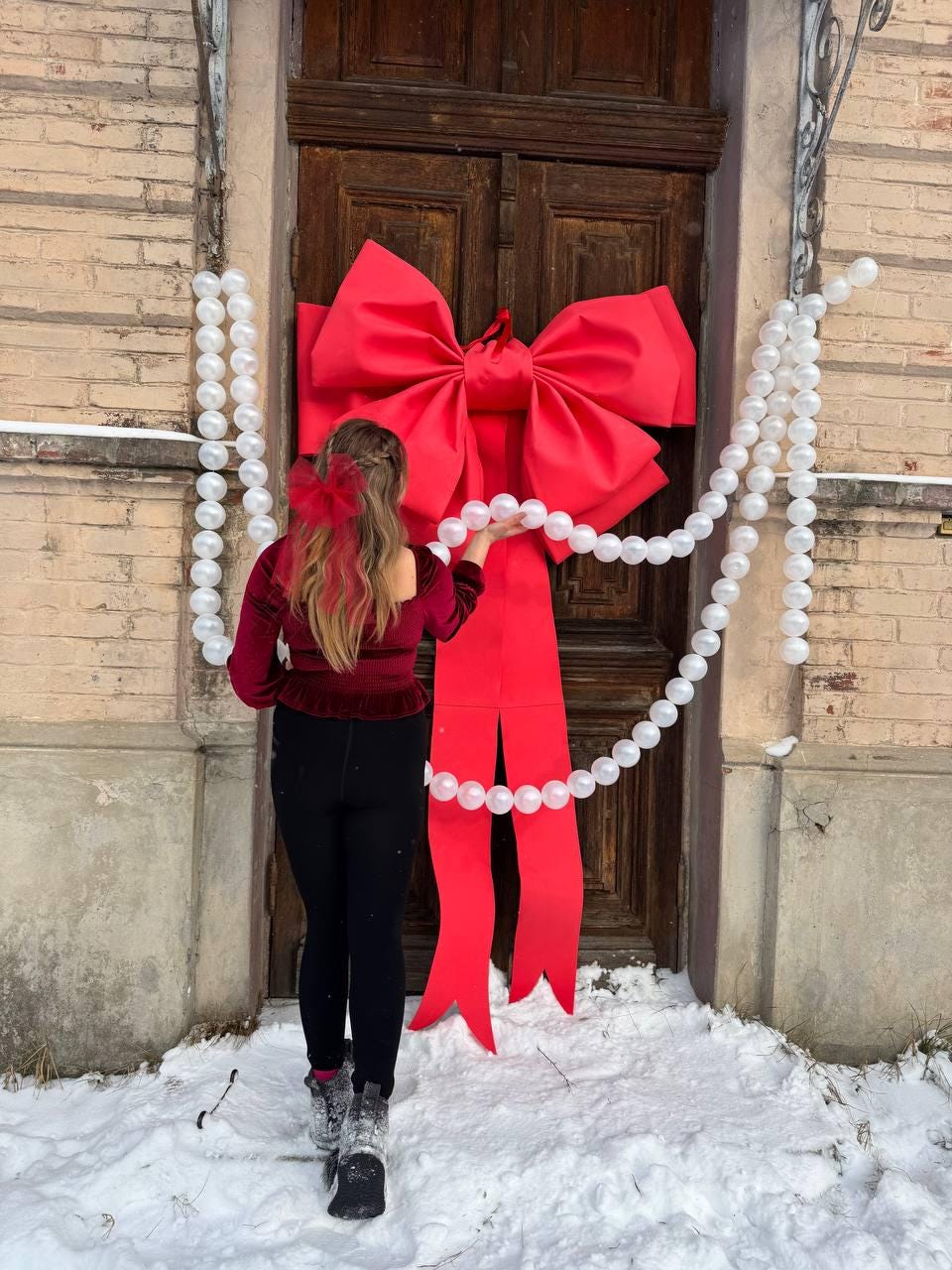 May include: A large, vibrant red bow adorns a wooden door, with long ribbon tails cascading down. White pearl-like strands hang on either side. The scene is set outdoors, with snow on the ground, suggesting a festive holiday theme.