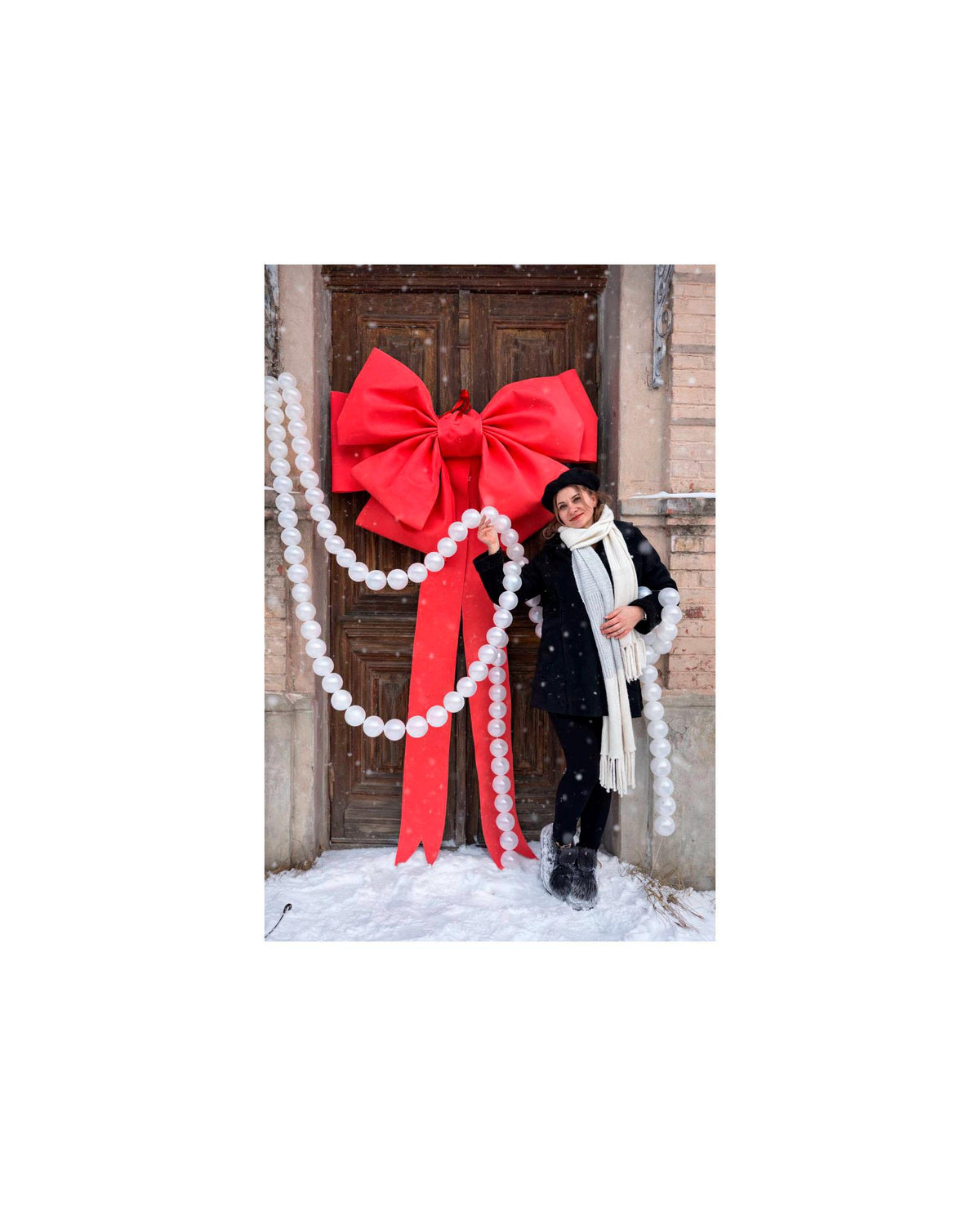 May include: A large red bow decorates a wooden door, with a string of white orbs draped nearby. The scene is set in a snowy environment, suggesting a festive holiday theme. The bow is the focal point, adding a pop of color against the door's brown.