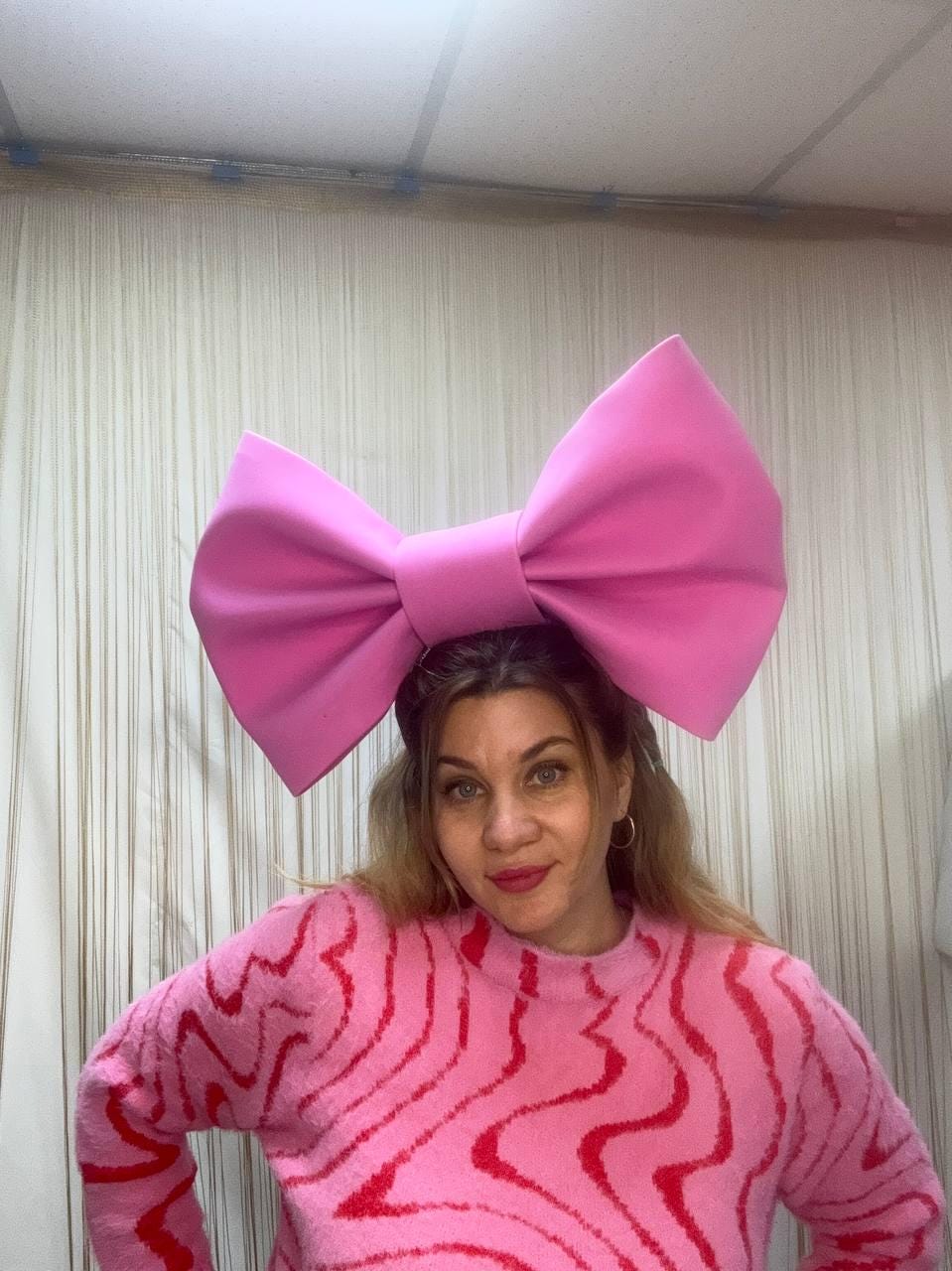 May include: A large, bright pink bow headband. The woman in the image is wearing a pink sweater with a red wavy pattern. The bow is the focal point, adding a bold, playful touch to the outfit. The background is a neutral color.