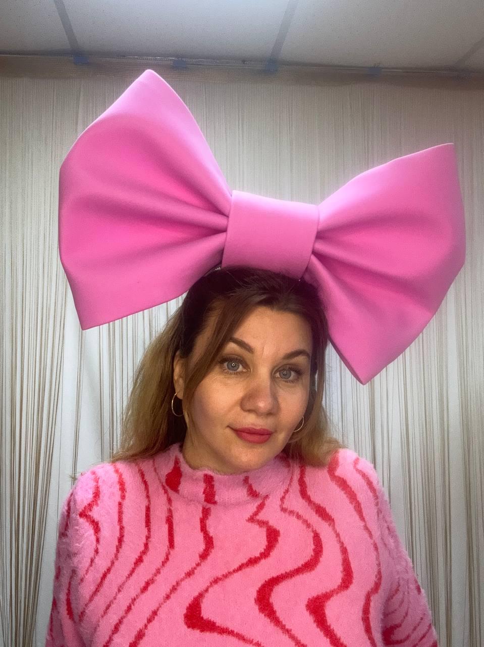 May include: A large, bright pink bow headband. The bow is a solid color and is worn on the head. The person in the image is wearing a pink sweater with a red wavy pattern.