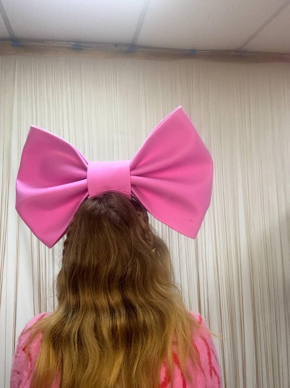 May include: A large, bright pink bow hair accessory. The bow is made of a smooth fabric and is positioned on the back of a person's head. The person is wearing a pink sweater. The background is a light-colored wall.