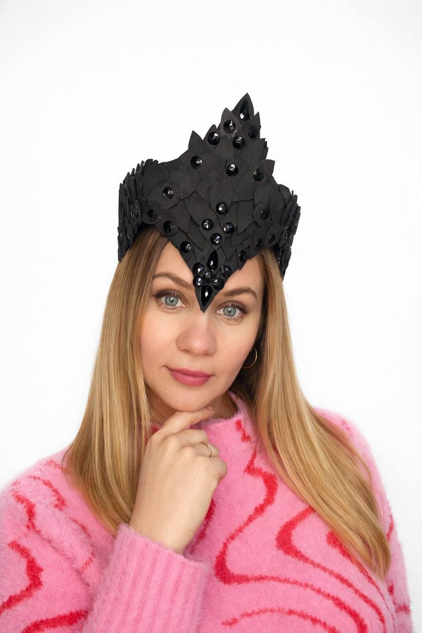 May include: A black crown with a pointed design and embellished with black gems. The crown is worn by a person wearing a pink sweater. The crown is a unique accessory.