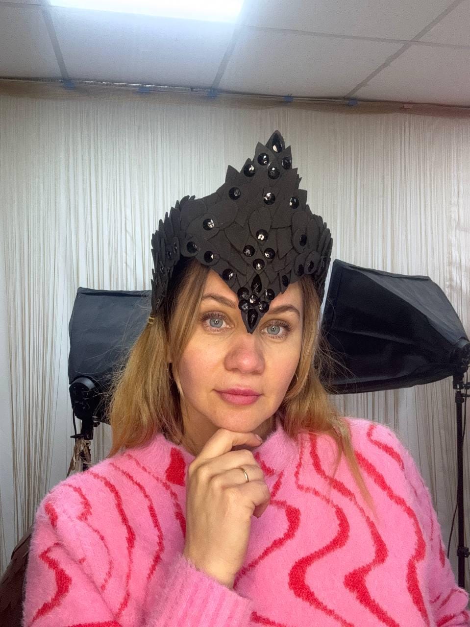 May include: A black crown with a pointed design and embellished with dark sequins. The crown is worn by a person wearing a pink sweater with a red wavy pattern. The background includes studio lighting and a white curtain.