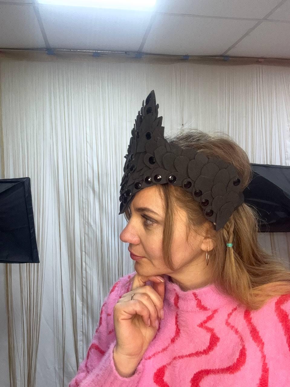 May include: A black crown with a spiky design and circular embellishments. The crown is worn on the head. The person is wearing a pink sweater with a red wavy pattern.
