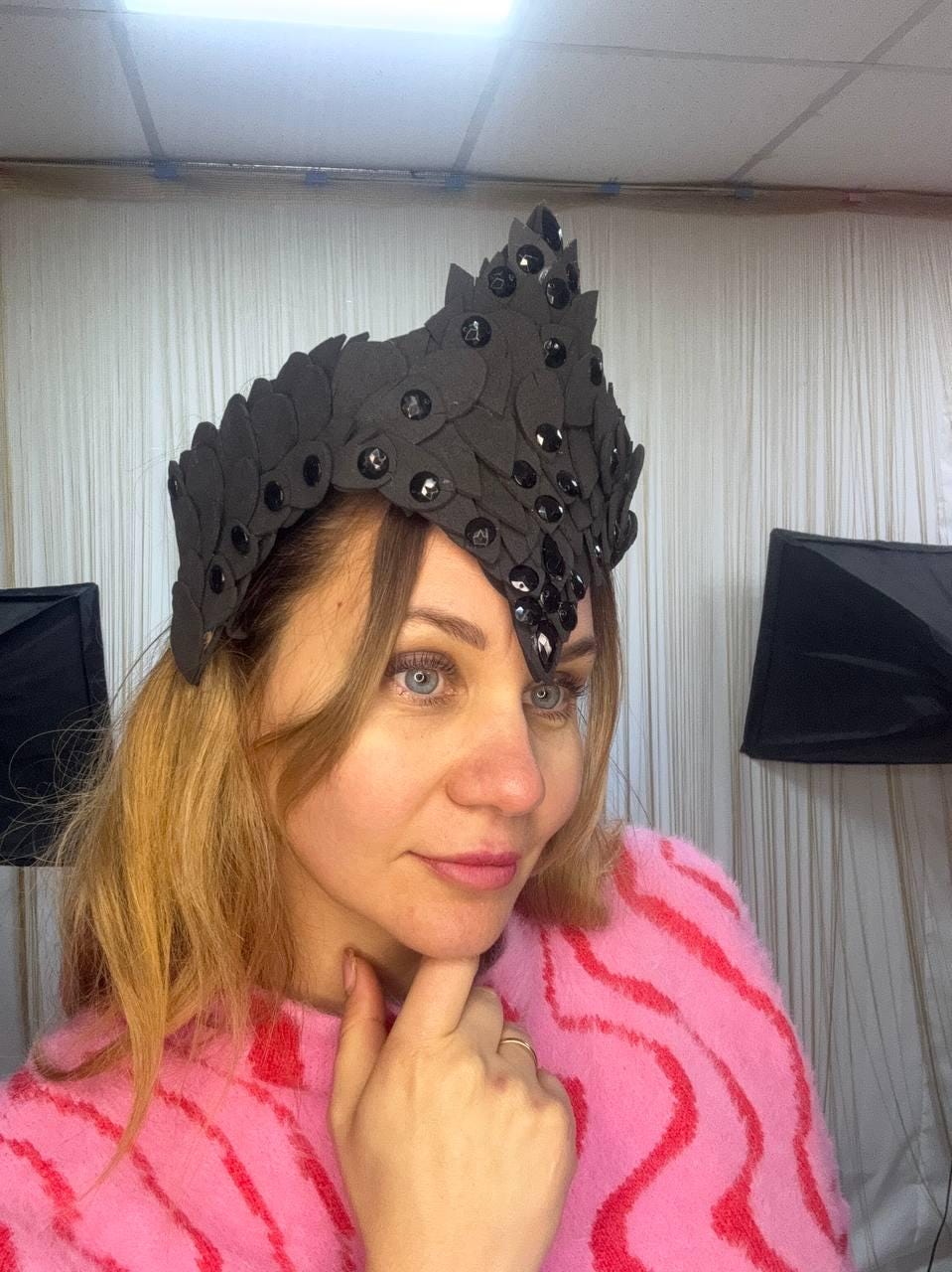 May include: A black crown-shaped headpiece with a leaf-like design and embellished with black gemstones. The headpiece is worn on the head. The person in the image is wearing a pink and red striped sweater.