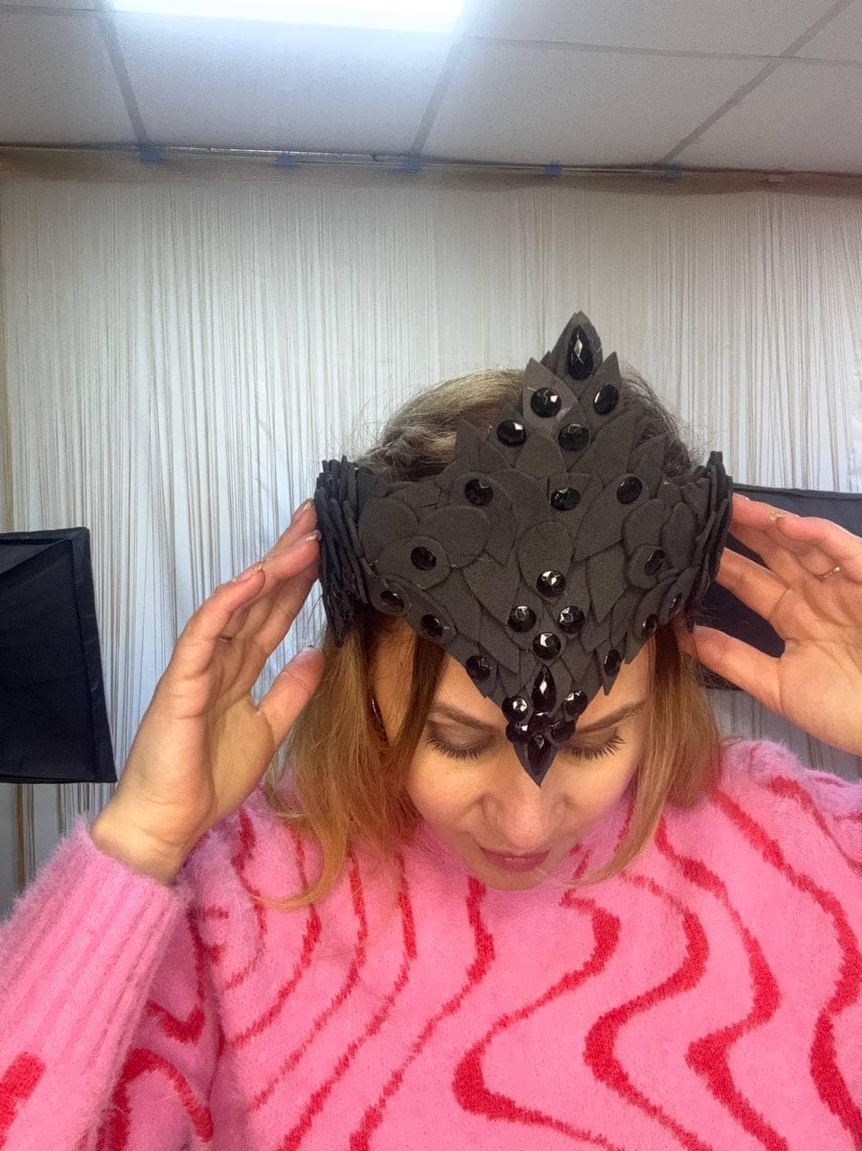 May include: A black crown with a pointed center and decorative embellishments. The crown is adorned with black gems and leaf-shaped details. The crown is being held by a person wearing a pink sweater.
