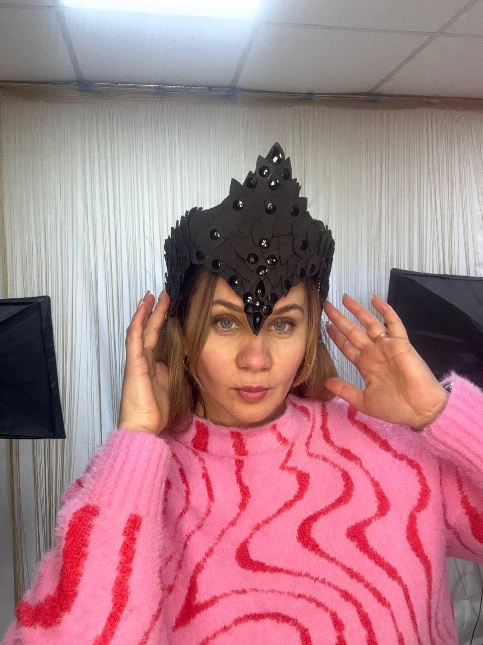 May include: A black crown with a pointed design and embellished with dark rhinestones. The crown is worn by a person wearing a pink sweater with a red wavy pattern.