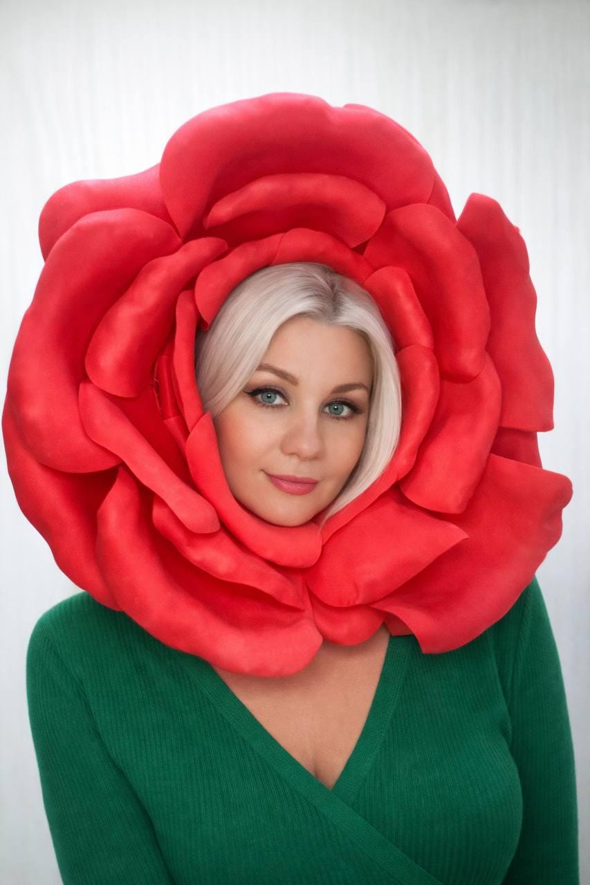 May include: A large, vibrant red rose headpiece. The petals frame the face of a person with blonde hair and green sweater. The headpiece is a striking costume element, perfect for themed events or artistic expression.
