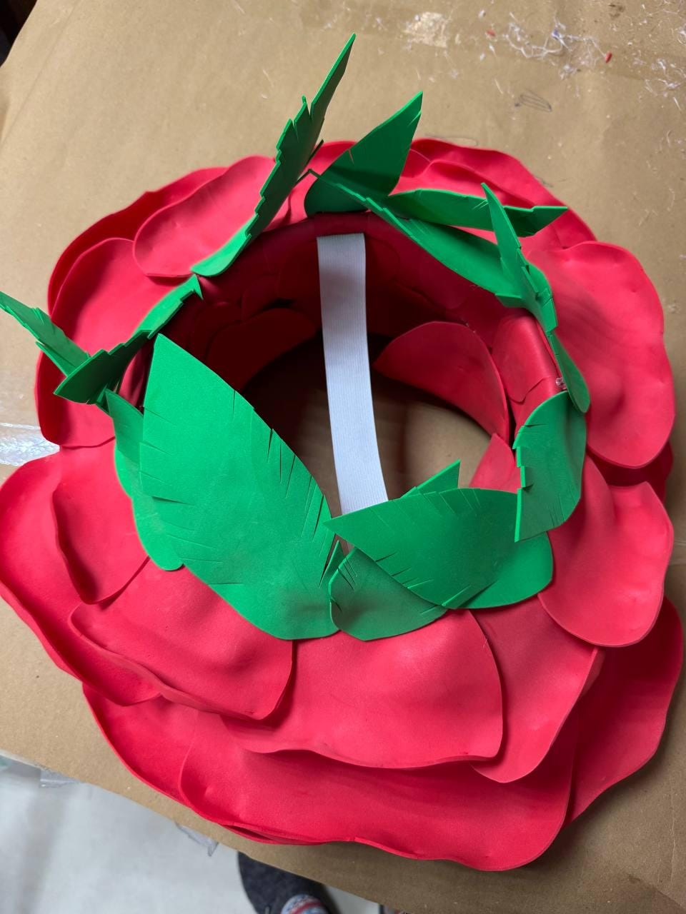 May include: A large, red rose-shaped headpiece with layered petals. The headpiece features green foam leaves around the inner rim and a white strap for securing. This costume accessory is ideal for themed events.