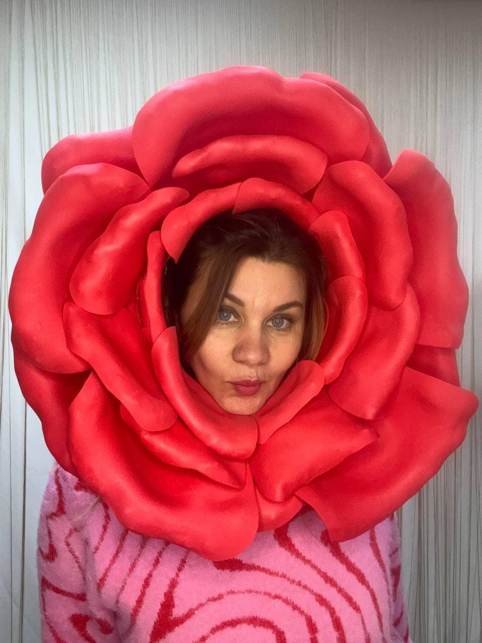 May include: A large, vibrant red rose costume worn by a person. The rose is the focal point, with detailed petals. The person is wearing a pink sweater with a wavy pattern. The background is a plain white wall.