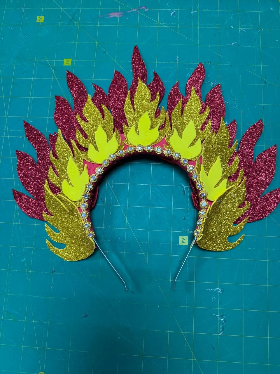 May include: A festive headpiece with a flame design. The crown features red, gold, and yellow glittery foam cutouts, with a row of small, clear rhinestones along the base. The headband is red, and the design is supported by thin, silver wire.