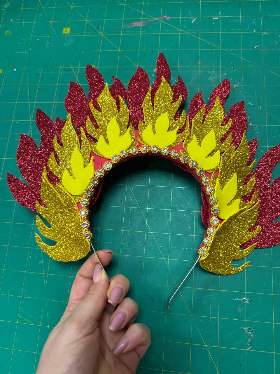 May include: A decorative headpiece with a flame design. The headpiece features red, yellow, and gold glittery foam cutouts, with a red base and a row of small, clear rhinestones. The headpiece is held by a thin wire.