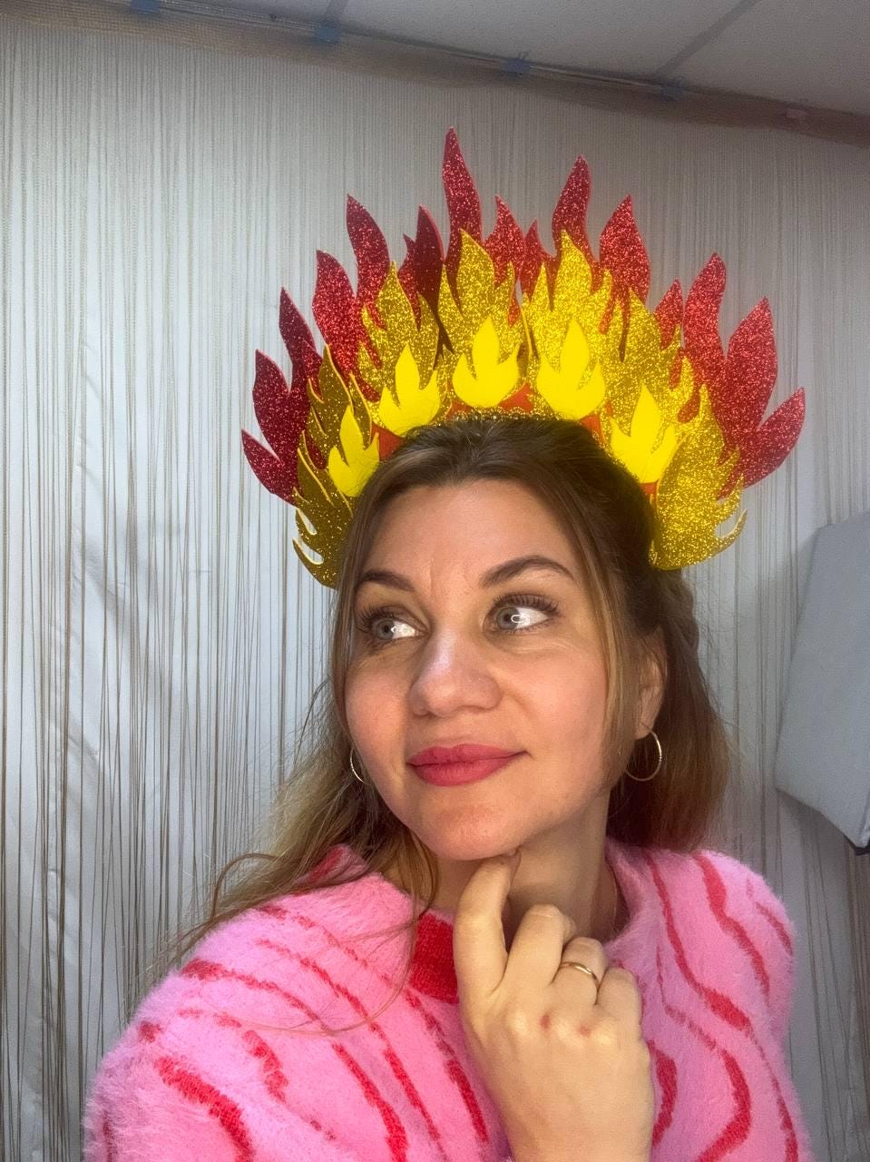 May include: A decorative headpiece resembling flames, featuring red, yellow, and gold glitter accents. The flame-shaped design is arranged in a crown shape. The headpiece is worn by a person wearing a pink sweater.