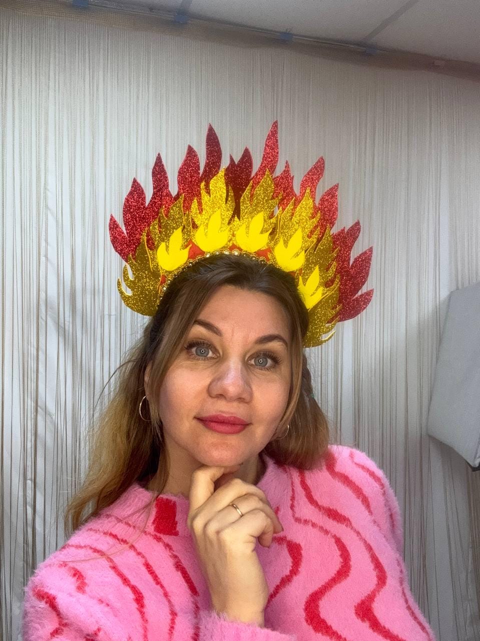 May include: A decorative headpiece resembling flames, featuring red and yellow glittery accents. The crown is worn by a person wearing a pink sweater. The headpiece is likely for a costume or themed event.