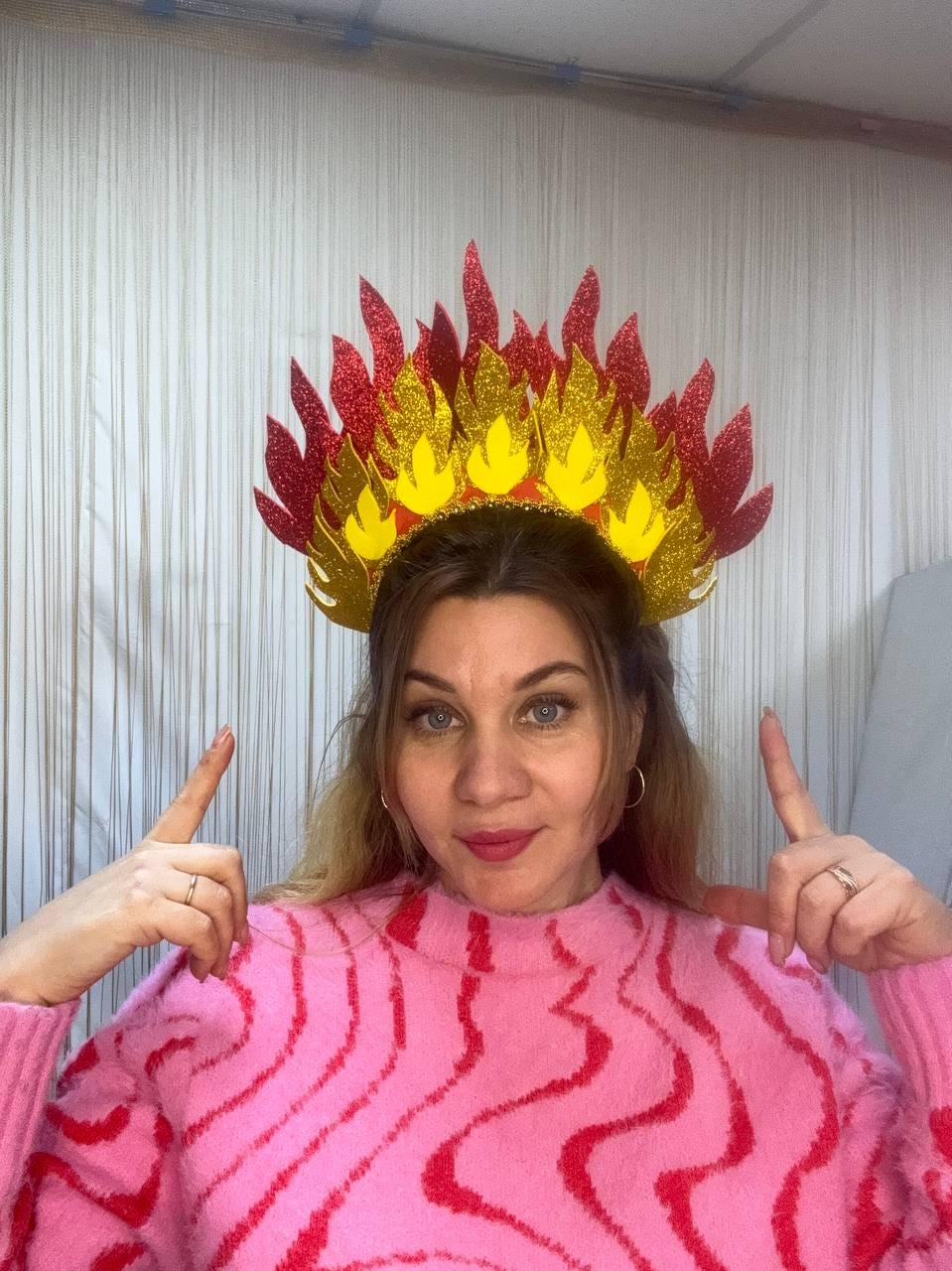 May include: A decorative headpiece resembling flames, featuring red, gold, and yellow glitter accents. The crown-like design is worn on the head. The person in the image is wearing a pink sweater with a red wavy pattern.