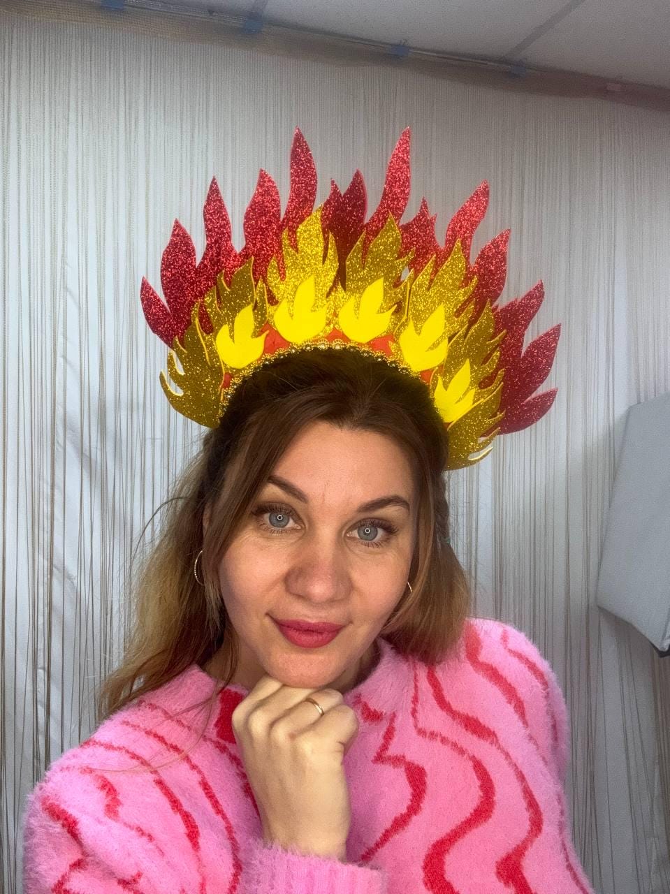 May include: A decorative headpiece resembling flames, featuring red, yellow, and gold glitter accents. The crown is designed with a flame-like pattern, perfect for themed events or costumes. The headpiece is worn by a person wearing a pink sweater.