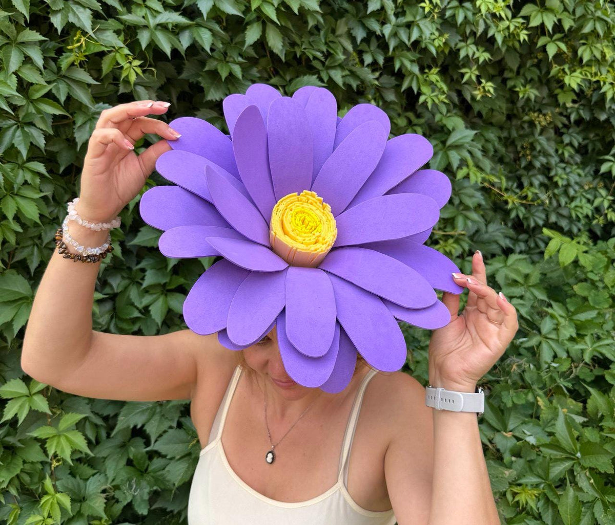 May include: A large, purple flower-shaped headpiece with a yellow center. The petals are a vibrant purple, and the center is a textured yellow. The headpiece is worn by a person in front of a green leafy background.