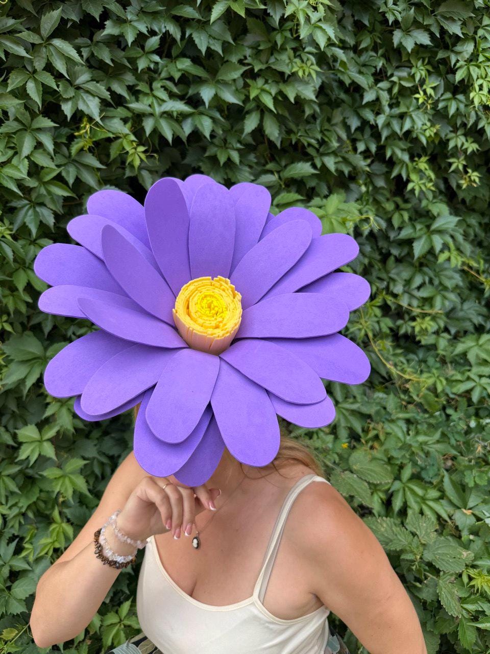 May include: A large, purple flower headpiece with a yellow center. The petals are layered and the headpiece is worn by a person. The background is a wall of green leaves.